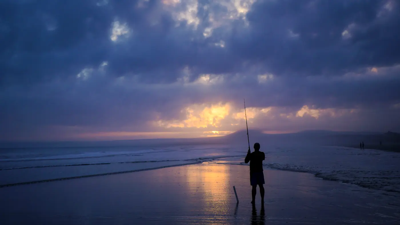 Man fishing from the beach at dusk under a cloudy sky