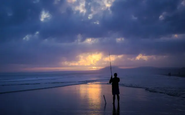 Man fishing from the beach at dusk under a cloudy sky