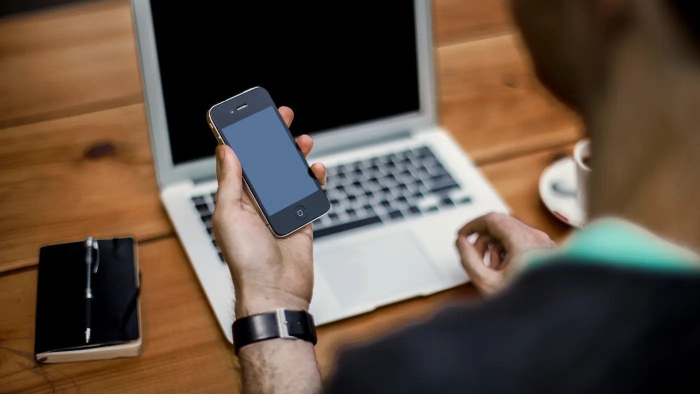Man checking his phone at a laptop on a wooden desk