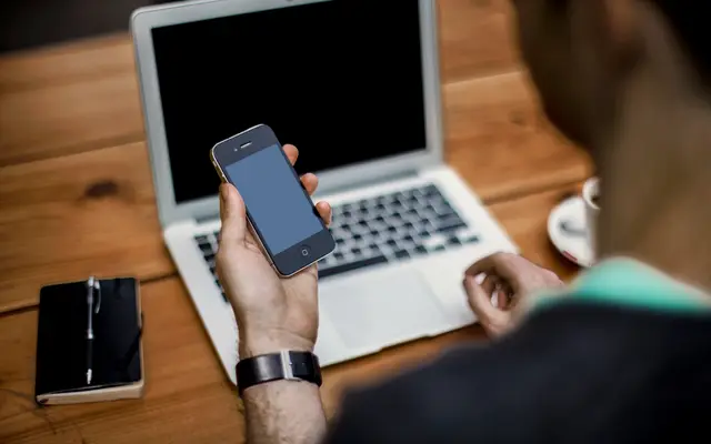 Man checking his phone at a laptop on a wooden desk