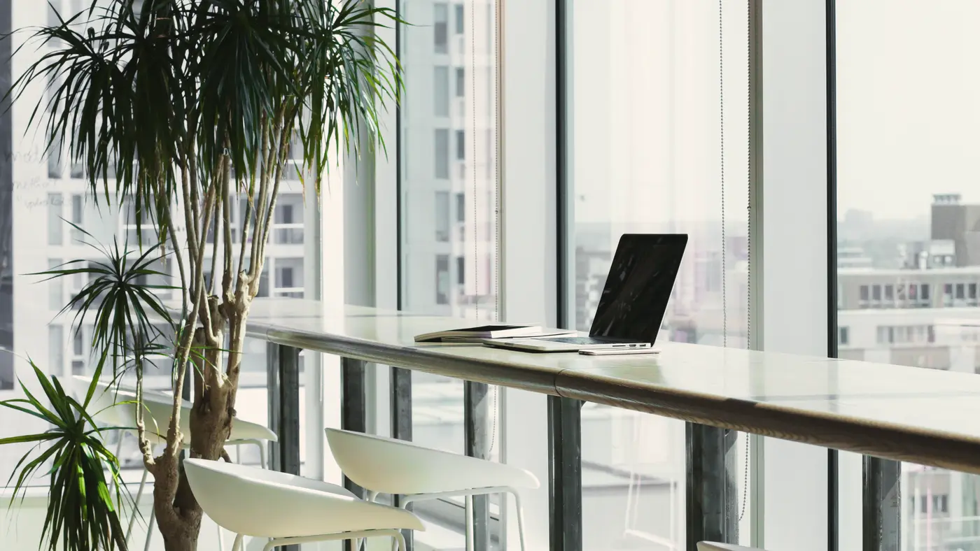 Laptop on a long desk by tall city windows with plants