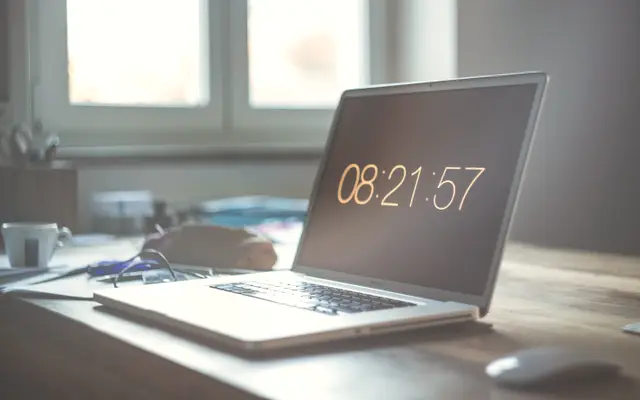 Laptop showing a digital clock on a desk by the window