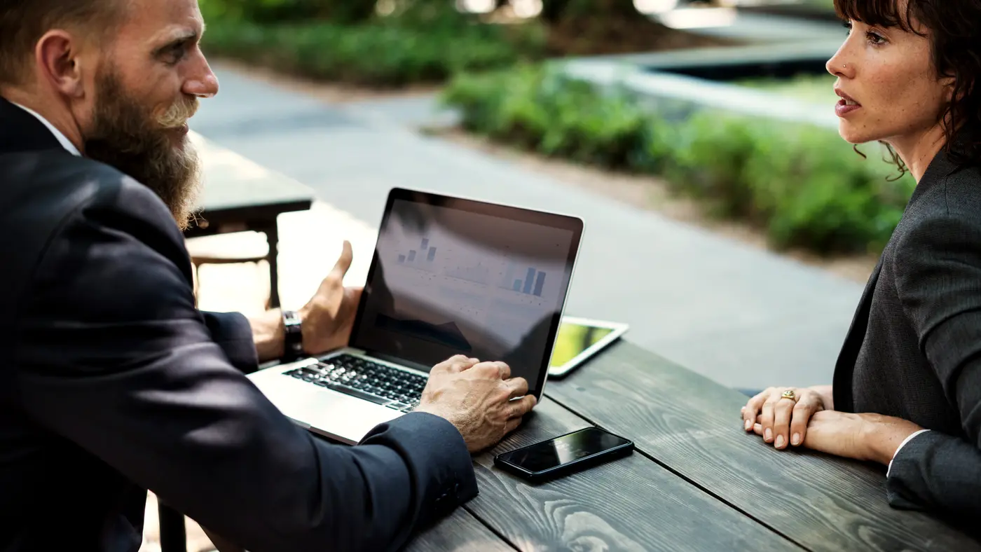 Two business people talking at an outdoor table over a laptop
