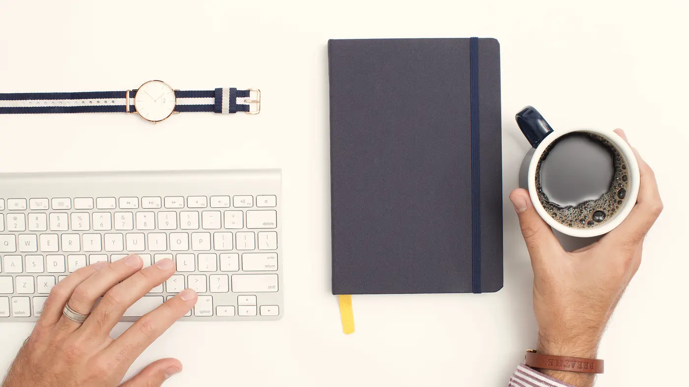 Hand on keyboard beside a notebook, watch and coffee cup