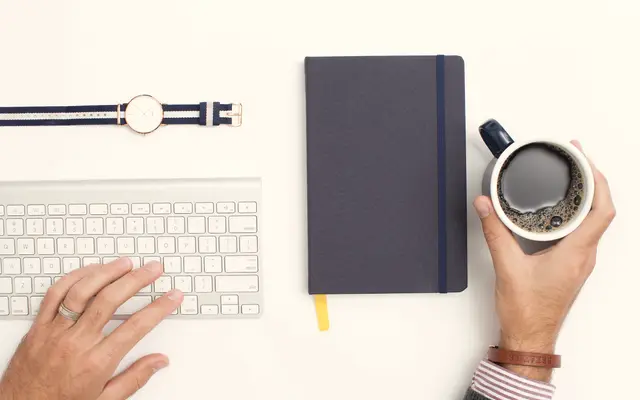 Hand on keyboard beside a notebook, watch and coffee cup