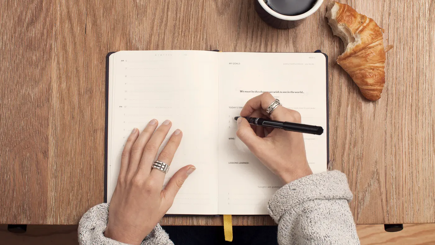 Hands writing in a planner beside coffee on a wooden table
