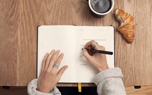 Hands writing in a planner beside coffee on a wooden table