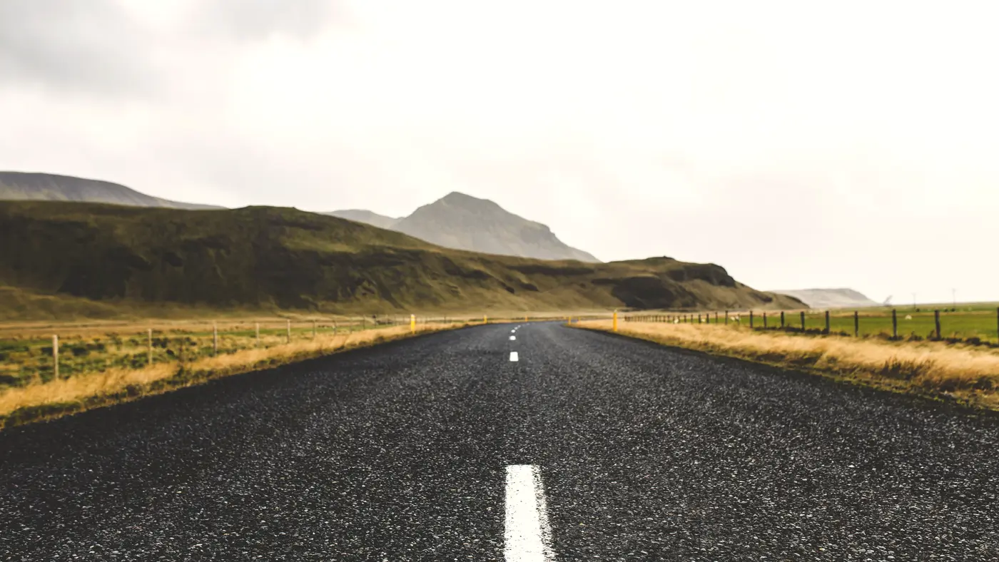 Empty asphalt road heading toward green hills under overcast sky