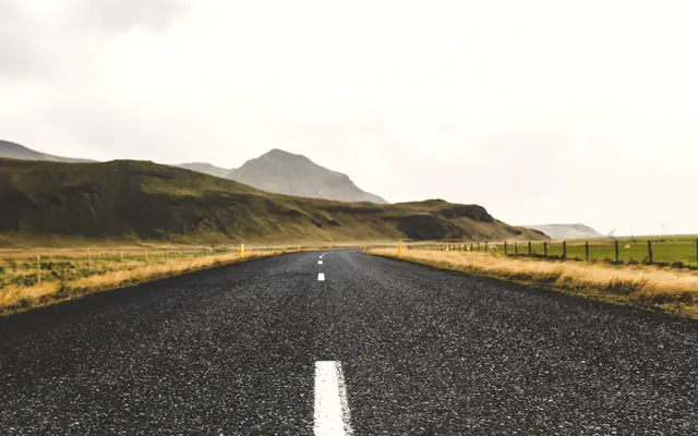 Empty asphalt road heading toward green hills under overcast sky
