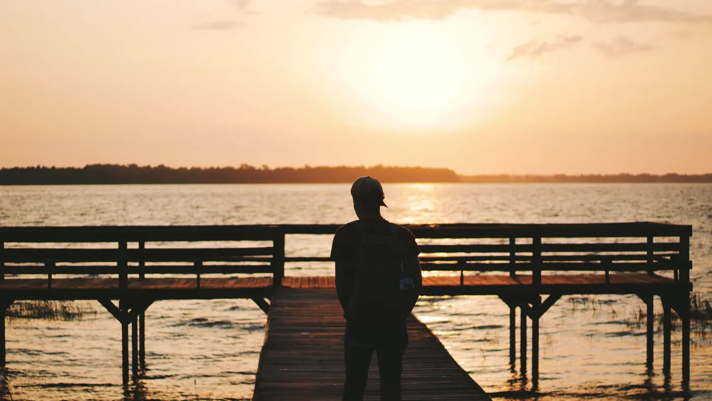 Man standing on a wooden pier facing a golden lake sunset
