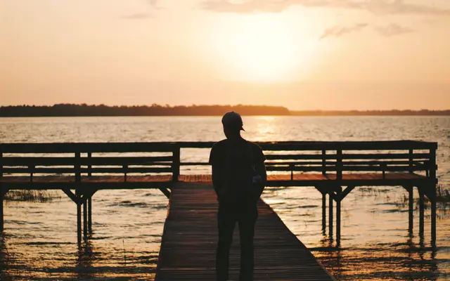 Man standing on a wooden pier facing a golden lake sunset