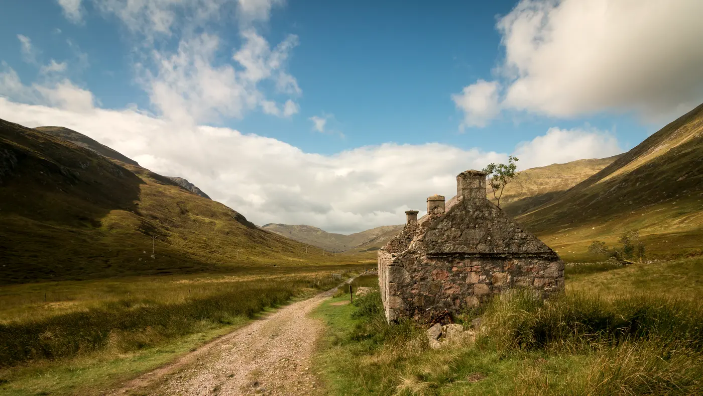 Dirt road winding through a green valley past a stone cottage