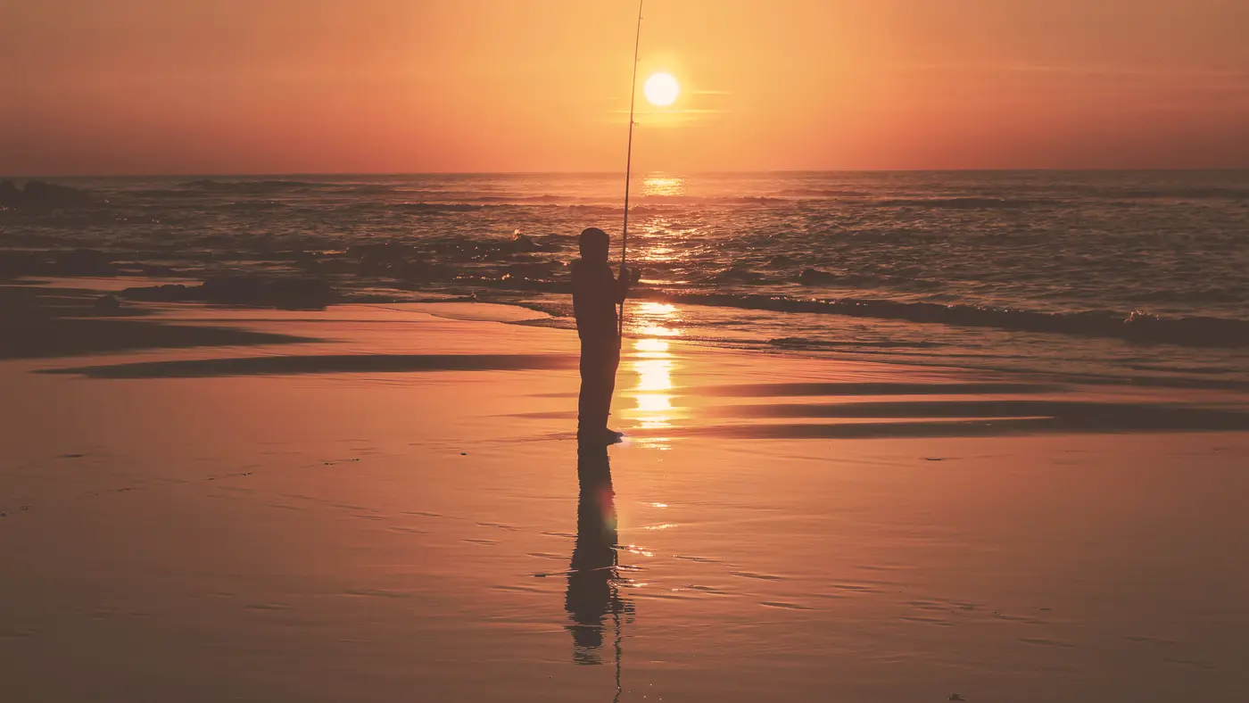 Angler casting into the surf at an orange sunset