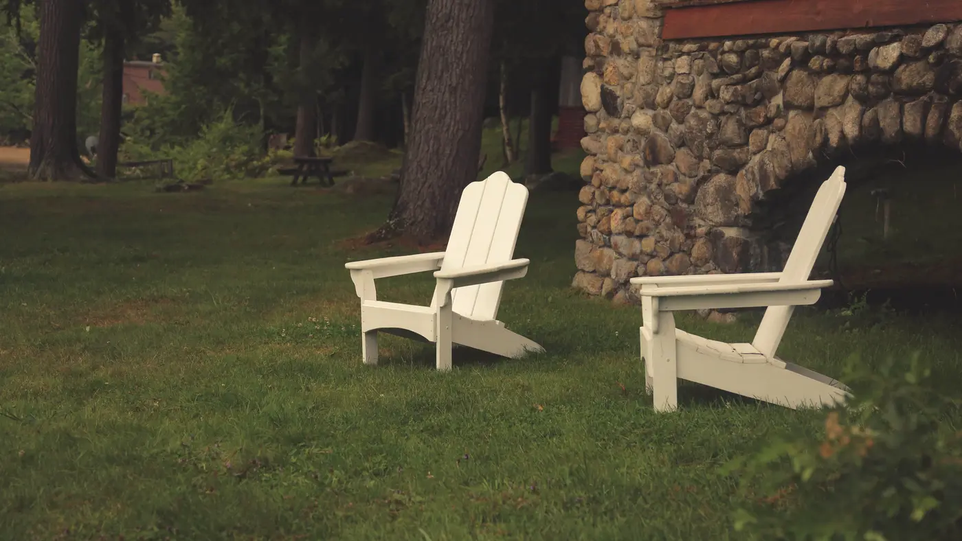 Two white Adirondack chairs on a lawn beside a stone house