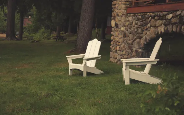 Two white Adirondack chairs on a lawn beside a stone house