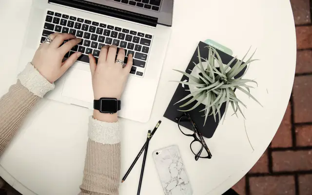 Hands typing on a laptop on a white table with notebook